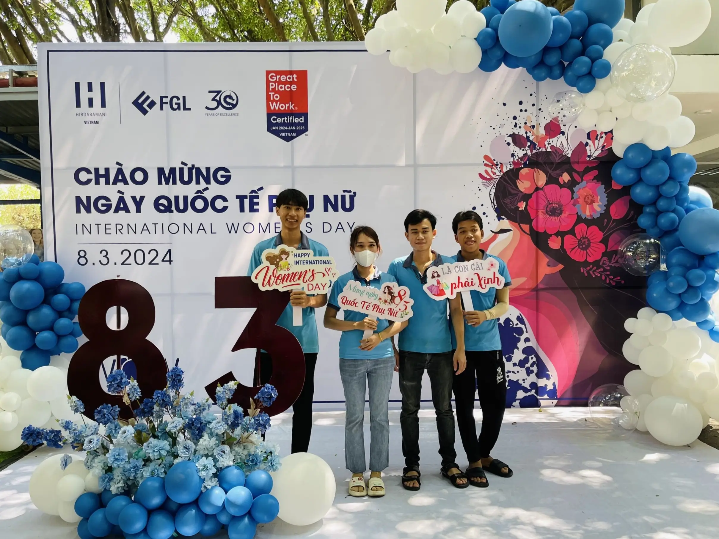Few employees holding tags and flowers in front of event banner for international women's day 